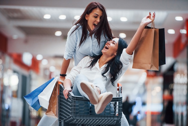 Foto dos mujeres jóvenes se divierten corriendo y montando en la cesta de la compra en el supermercado.