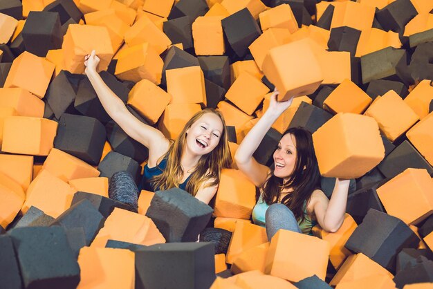 Dos mujeres jóvenes divirtiéndose con bloques blandos en el interior del parque infantil en el foso de gomaespuma en el centro de trampolín