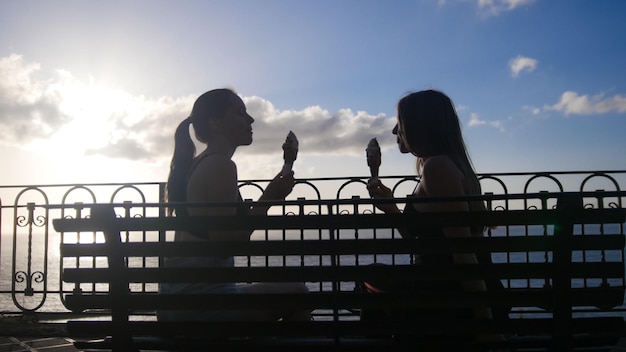 Dos mujeres jóvenes están sentadas con el helado en el banco frente al mar