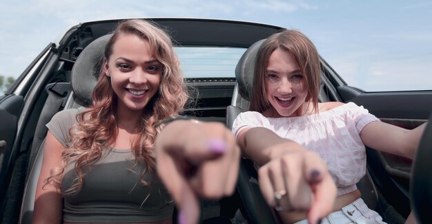 Dos mujeres jóvenes felices sentadas en un auto y señalándote