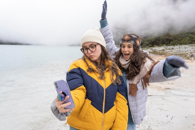 Dos mujeres jóvenes gesticulando divertidamente mientras se toman un selfie en una laguna en invierno