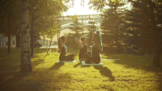 Dos mujeres jóvenes y un hombre haciendo yoga en el parque bajo los rayos del sol vespertino