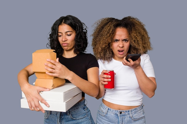 Dos mujeres jóvenes, una latina y otra con cabello afro, se ríen mientras sostienen pizzas y hamburguesas.