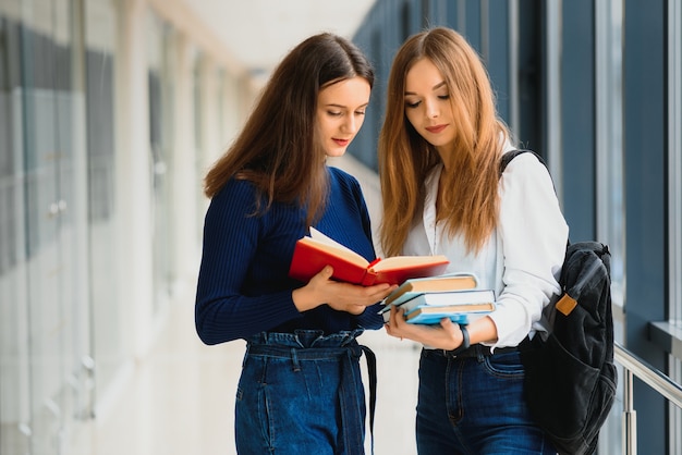 Dos mujeres jóvenes con libro charlando mientras está de pie en el pasillo de la universidad