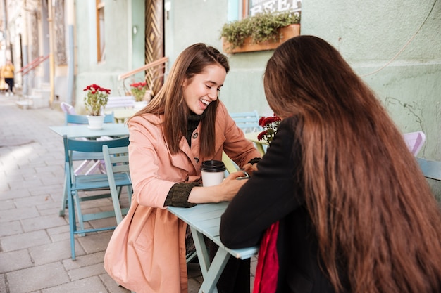 Dos mujeres jóvenes lindas alegres bebiendo café y riendo en la cafetería al aire libre