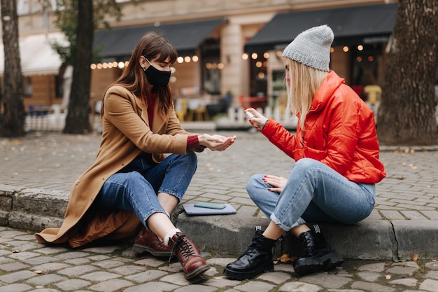 Dos mujeres jóvenes con máscaras médicas pasando tiempo libre juntas al aire libre y usando antiséptico para las manos. Prevención de la propagación del coronavirus.