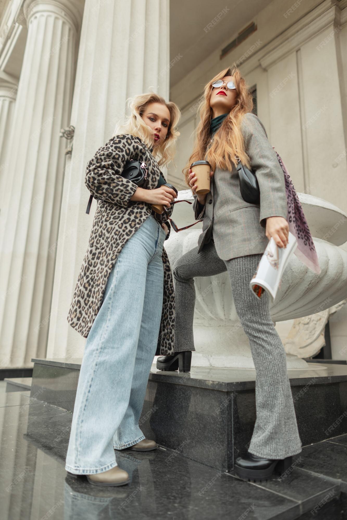 Dos mujeres de moda con una taza de café y una revista con ropa elegante de otoño con de leopardo, pantalones vaqueros, traje elegante y puestos de zapatos y poses