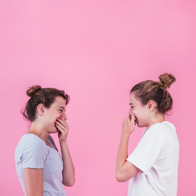 Dos mujeres jóvenes de pie cara a cara riendo con la boca tapada sobre fondo rosa