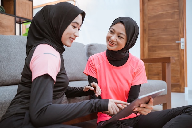 Dos mujeres jóvenes con ropa deportiva hijab sonriendo mientras se relaja en el suelo mientras usa una tableta digital apoyado en el sofá en casa