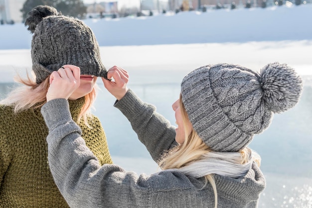 dos mujeres juegan a ponerse el sombrero sobre los ojos, de pie junto al río en invierno