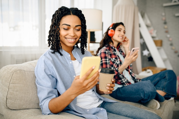 Dos mujeres lindas en auriculares escuchando música y bebiendo café en el sofá. Bonitas novias en auriculares se relajan en la habitación, amantes del sonido descansando en el sofá, persona de sexo femenino mediante teléfono móvil