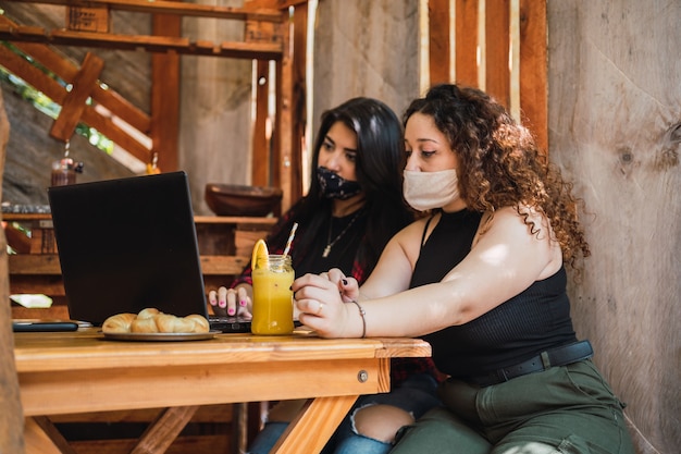 Dos mujeres con máscaras protectoras sentadas en un bar trabajando con una computadora portátil - Mujeres sentadas en un bar disfrutando de un jugo.