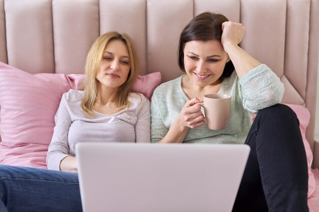 Dos mujeres de mediana edad sentadas en casa en la cama con una laptop