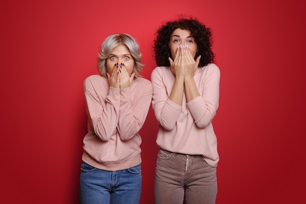 Dos mujeres mirando a la cámara con ojos de asombro tapándose la boca con las palmas de las manos rojo backgro ...