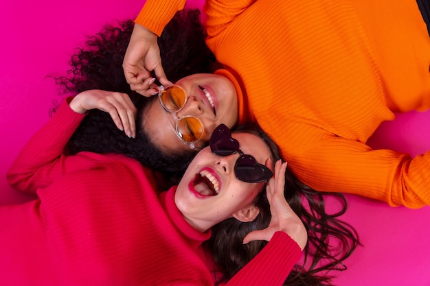 Dos mujeres multiétnicas con gafas de sol acostadas sonriendo en un estilo de vida de fondo rosa