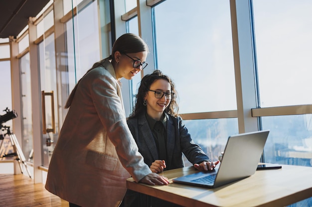 Dos mujeres de negocios buscan algo en una computadora portátil Concepto de cooperación empresarial y trabajo en equipo Mujeres jóvenes sonrientes en el escritorio de la oficina Gente exitosa moderna