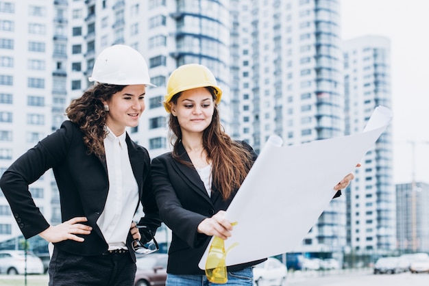 Dos mujeres de negocios en los cascos de protección y gafas de seguridad en busca de esquemas de construcción, concepto arquitectónico