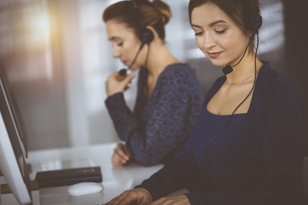 Dos mujeres de negocios conversan con los clientes a través de auriculares, mientras están sentadas en el escritorio de una oficina moderna y soleada. Grupo diverso de personas en un call center. Telemercadeo y atención al cliente.