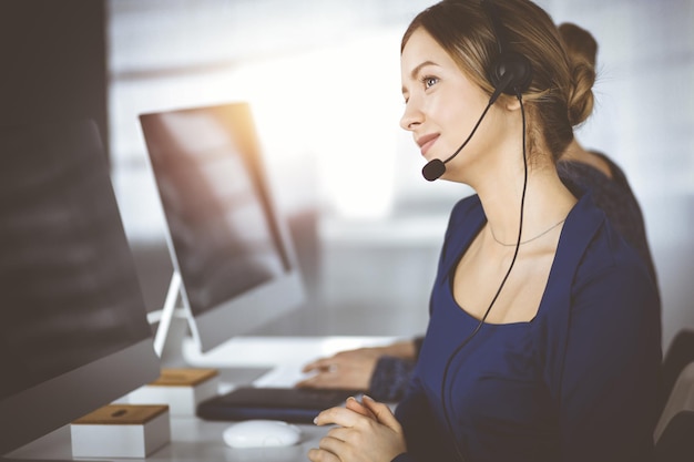 Dos mujeres de negocios conversan con los clientes a través de auriculares, mientras están sentadas en el escritorio de una oficina moderna y soleada. Grupo diverso de personas en un call center. Telemercadeo y atención al cliente.
