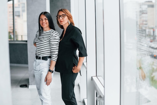 Dos mujeres de negocios juntas en el interior de la oficina cerca de una ventana grande