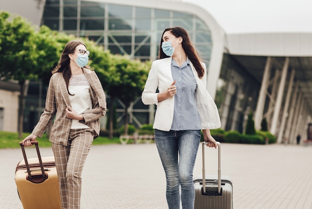 Dos mujeres de negocios en máscaras protectoras con maletas cerca del aeropuerto