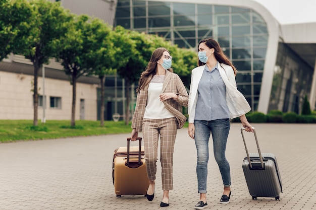 Dos mujeres de negocios en máscaras protectoras con maletas van al aeropuerto