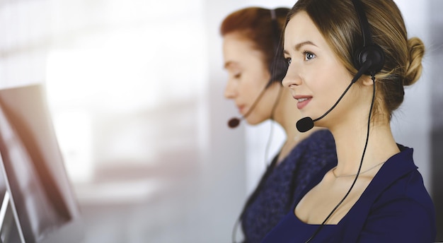 Dos mujeres de negocios tienen conversaciones con los clientes a través de auriculares, mientras están sentadas en el escritorio de una oficina moderna y soleada. Grupo de personas diversas en un centro de llamadas. Telemarketing y atención al cliente