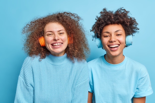 Dos mujeres optimistas y guapas con cabello rizado sonríen ampliamente y se divierten mientras escuchan música a través de auriculares y disfrutan de su pista favorita vestidas casualmente aisladas sobre una pared azul. Concepto de estilo de vida