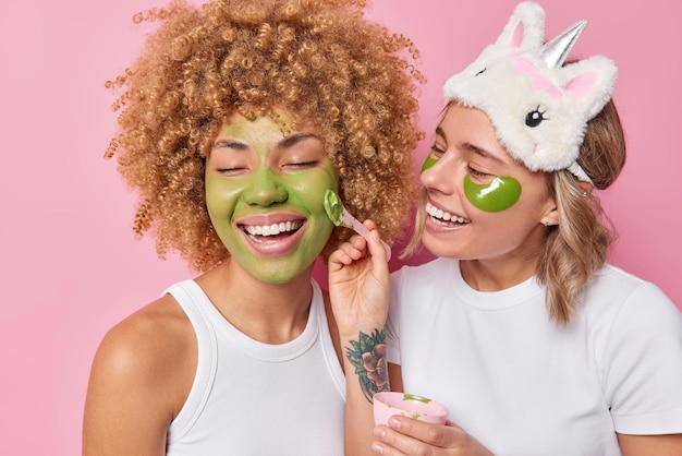 Dos mujeres positivas se someten a procedimientos de belleza, aplican una máscara hidratante verde y los parches cuidan la piel, usan camisetas blancas informales que se encuentran muy aisladas sobre fondo rosa Concepto de cuidado facial