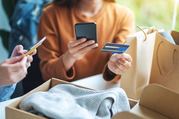 Dos mujeres que usan teléfono móvil y tarjeta de crédito para comprar en línea con una bolsa de compras y una caja postal de ropa en la mesa