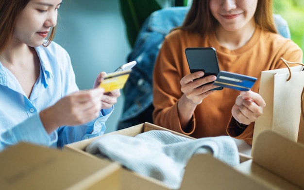 Dos mujeres que usan teléfono móvil y tarjeta de crédito para comprar en línea con una bolsa de compras y una caja postal de ropa en la mesa