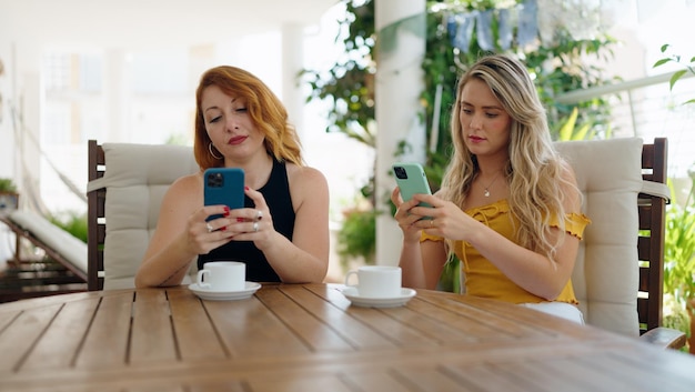 Dos mujeres que usan teléfonos inteligentes y beben café sentadas en la mesa en la terraza de su casa