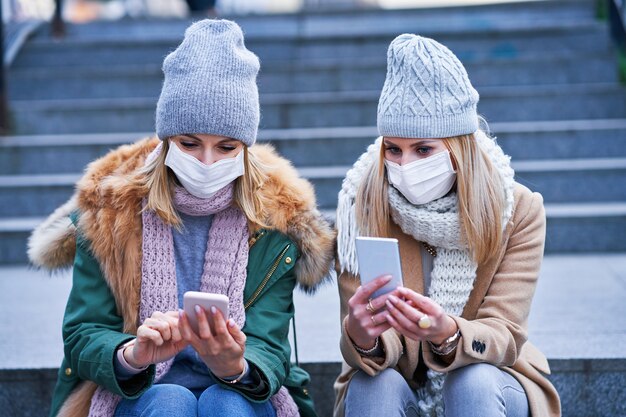 dos mujeres que usan teléfonos inteligentes y máscaras en la ciudad