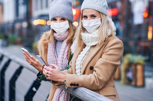 dos mujeres que usan teléfonos inteligentes y máscaras en la ciudad