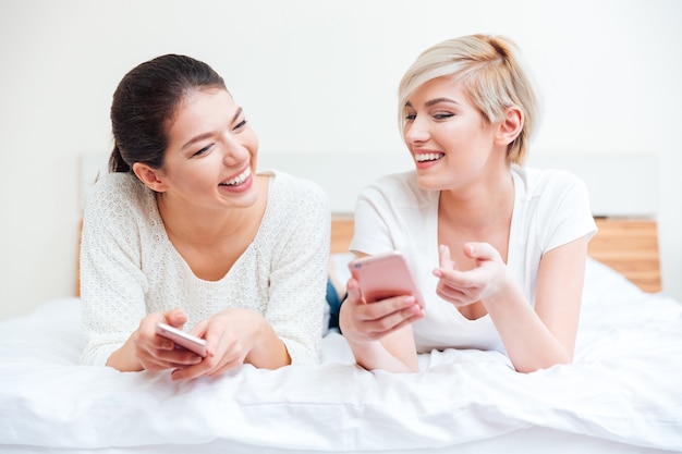 Dos mujeres riendo con teléfonos inteligentes en la cama en casa