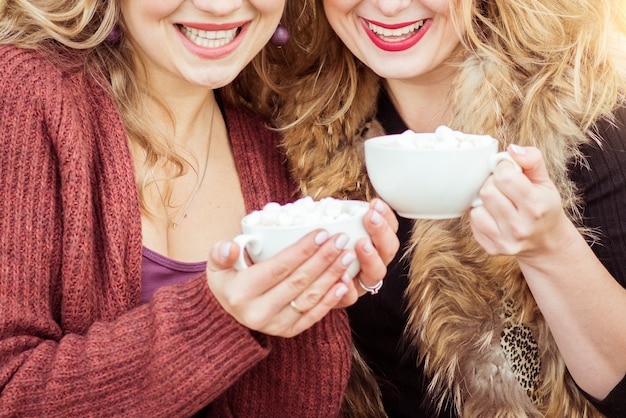 Dos mujeres sonrientes están sentados afuera con tazas de malvaviscos de chocolate caliente