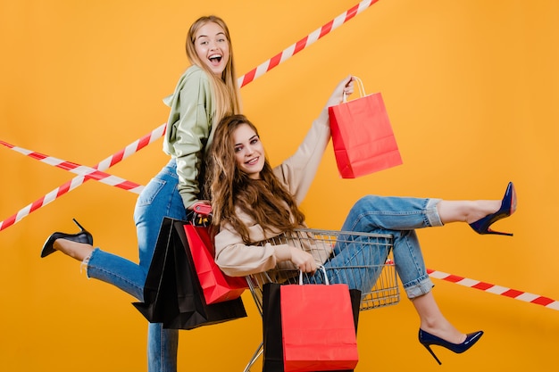 Dos mujeres sonrientes felices tienen carro con coloridos bolsos de compras y cinta de señal aislada sobre amarillo