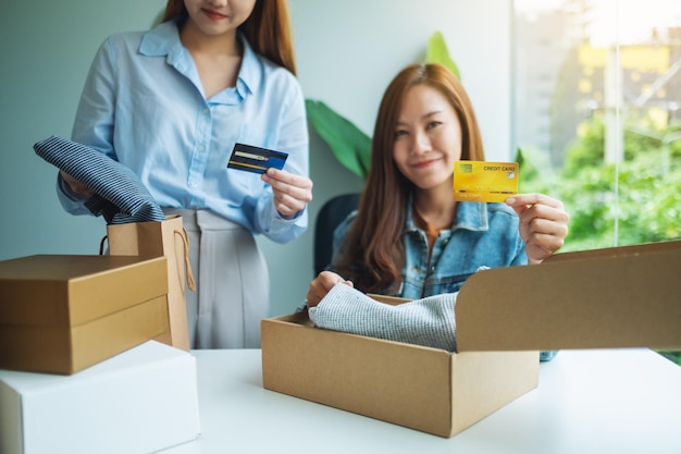 Dos mujeres sosteniendo y usando tarjeta de crédito para compras en línea mientras abren una caja de paquetes postales de ropa en casa