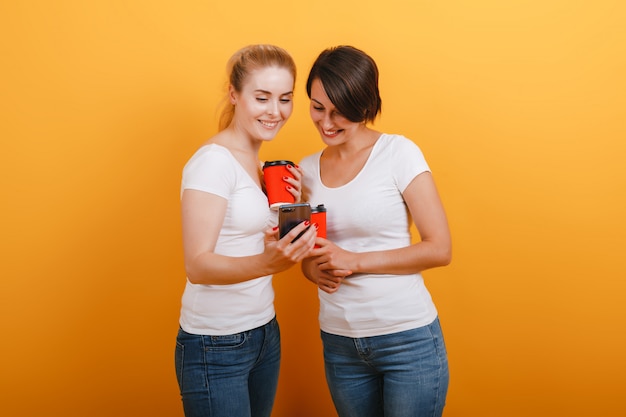 Dos mujeres sosteniendo un vaso de café de papel y mirando a la pantalla del teléfono inteligente contra el fondo amarillo del estudio