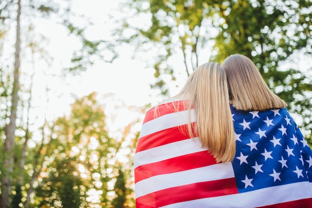 Dos mujeres sostienen la bandera de Estados Unidos y mirando hacia adelante. Vista trasera.