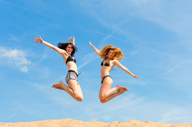 Dos mujeres en traje de baño felizmente saltando por encima de la playa de arena