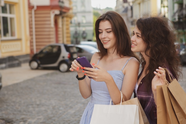 Dos mujeres usando un teléfono inteligente, caminando por la ciudad después de comprar juntas