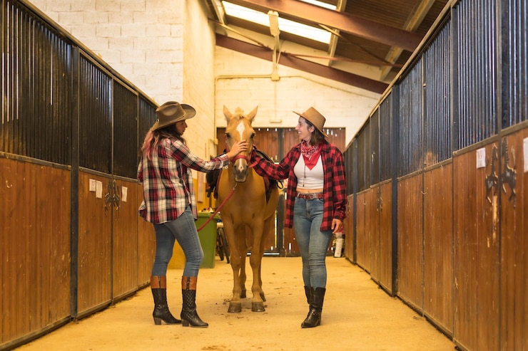 Foto dos mujeres vaqueras divirtiéndose y riendo con un caballo en un establo, sombreros de vaquero americano, camisas a cuadros y jeans