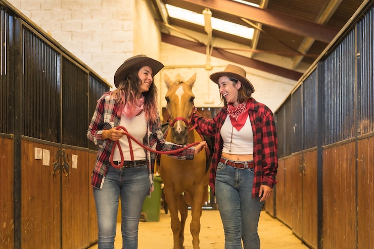 Foto dos mujeres vaqueras divirtiéndose y riendo con un caballo en un establo, con trajes de américa del sur