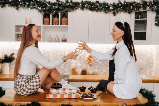 Dos niñas en un acogedor ambiente hogareño en la cocina con champán en sus manos para Navidad. Chicas sonrientes beben champán en una noche festiva