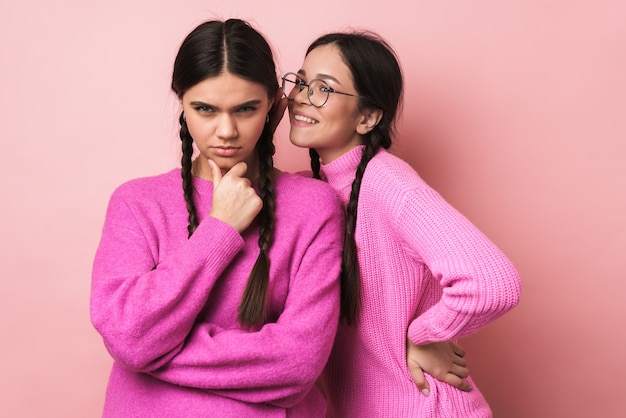 Dos niñas adolescentes felices con trenzas en ropa casual chismeando juntos aislado sobre pared rosa