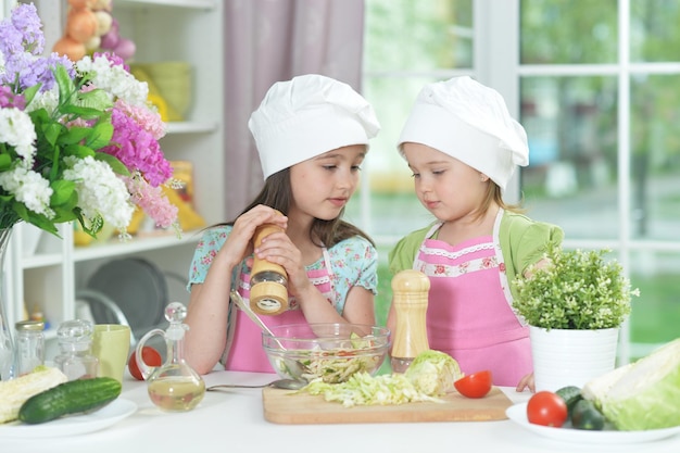 Dos niñas adorables con delantales preparando una deliciosa ensalada con repollo en la mesa de la cocina