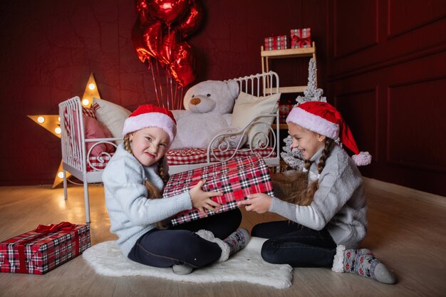 Dos niñas con coletas en gorras rojas de Navidad con cajas de regalo se sientan en el suelo cerca de la cama.