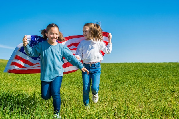 Dos niñas corriendo por un prado verde con un cielo azul, mostrando la bandera de los Estados Unidos. EE.UU.