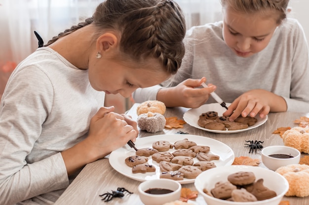 Dos niñas decoran galletas de jengibre de halloween en platos con glaseado de chocolate. Cocinar golosinas para la celebración de halloween. Estilo de vida